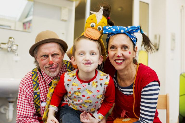 Zwei Klinikclowns und ein Kind posieren lachend in farbenfrohen Kostümen und bringen Freude in den Klinikalltag.