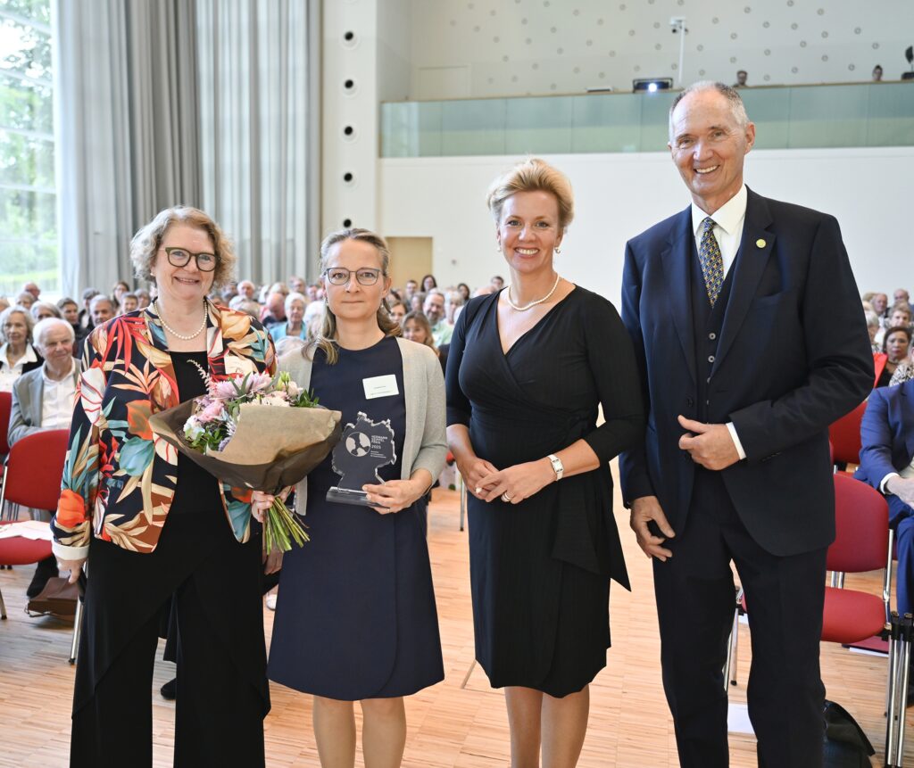 Vier Personen auf einer Bühne, eine Frau hält Blumen, eine weitere eine Glastrophäe, Publikum im Hintergrund.