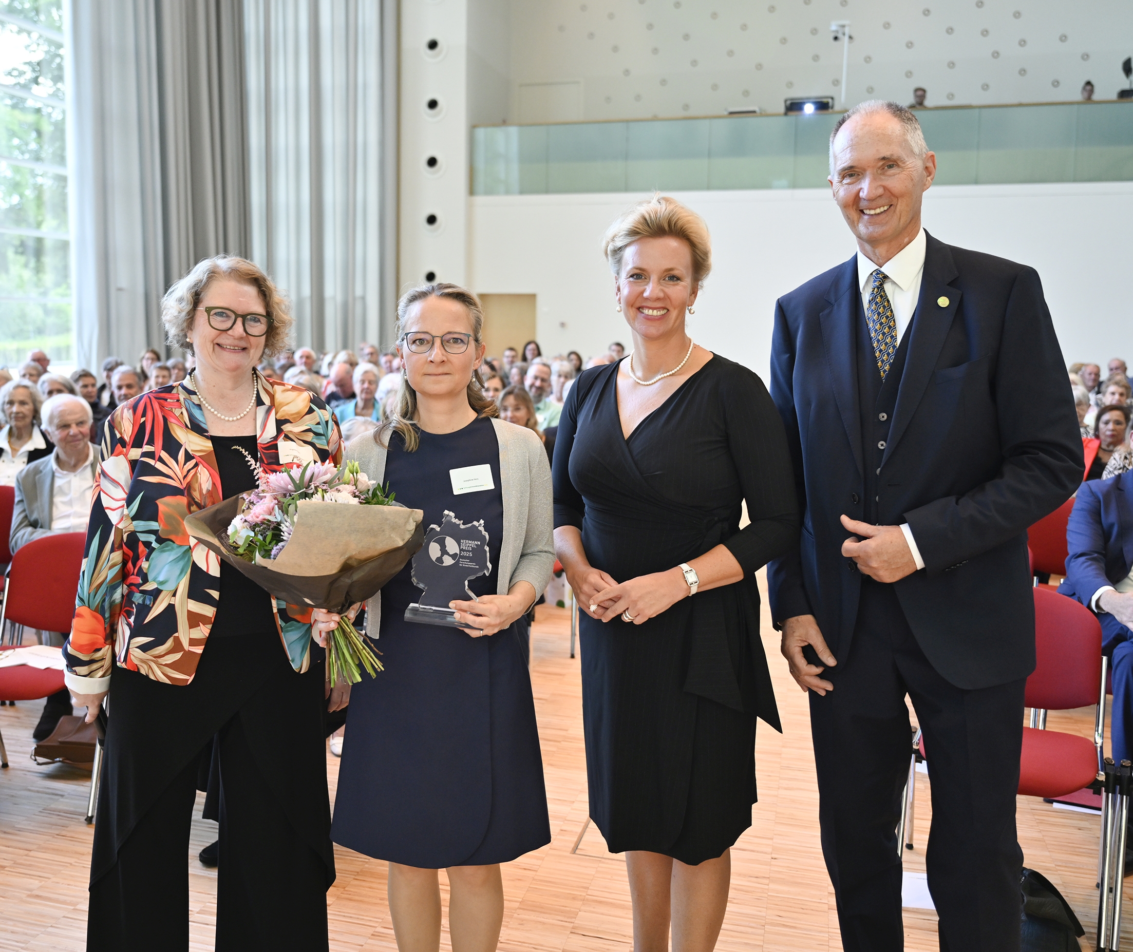 Vier Personen auf einer Bühne, eine Frau hält Blumen, eine weitere eine Glastrophäe, Publikum im Hintergrund.